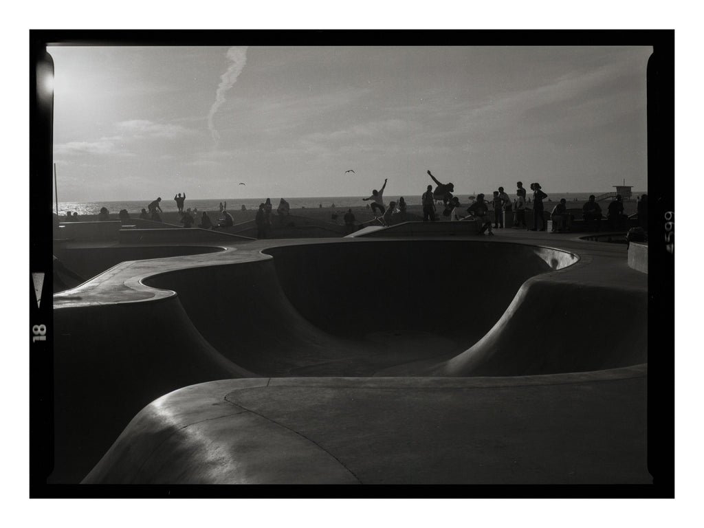 Black and white photograph of a skate park with people around a bowl at sunset in Venice beach california