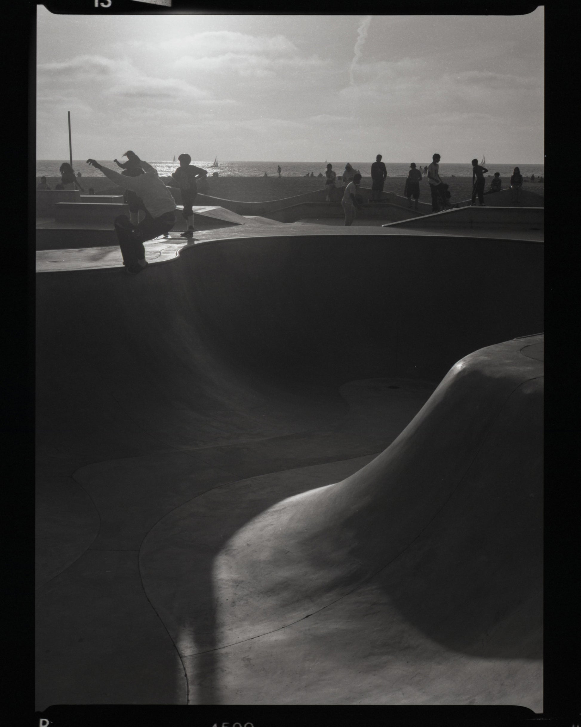 Black and white photo of a skateboarder at a venice beach skate park with people in the background