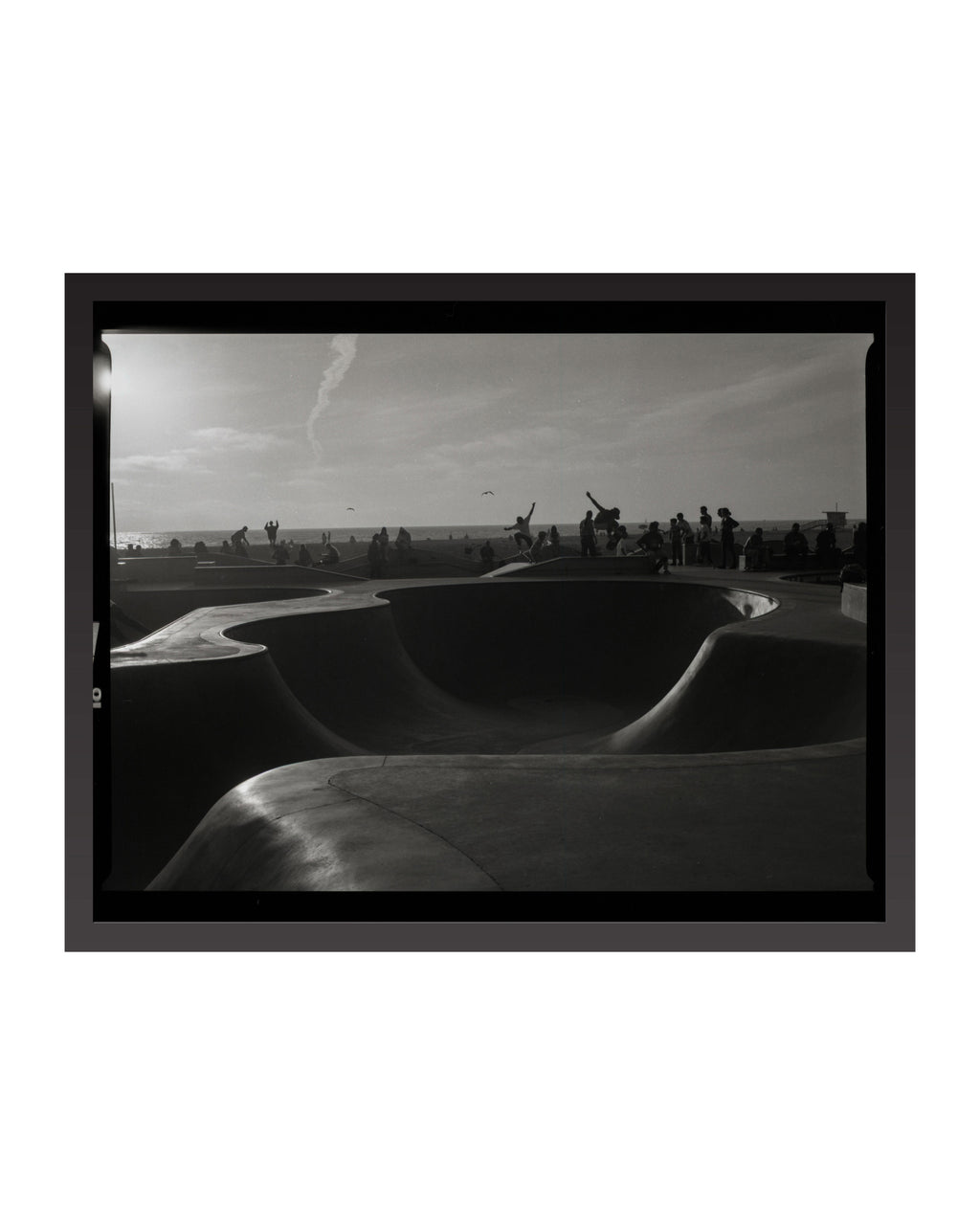 Black and white photograph of a skate park with a large bowl and people in the background.