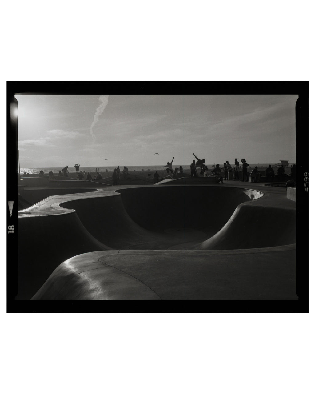 Black and white photograph of a venice beach skate park with people and a sunset.